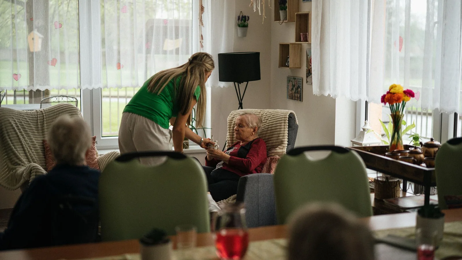 Une professionnelle sert une boisson à une résidente dans le salon lumineux d’un établissement pour personnes âgées