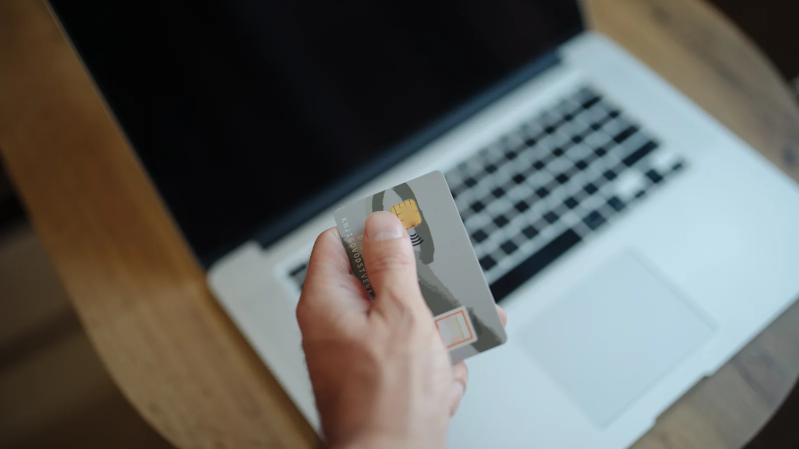 Une personne tient une carte bancaire devant un ordinateur portable ouvert sur une table en bois
