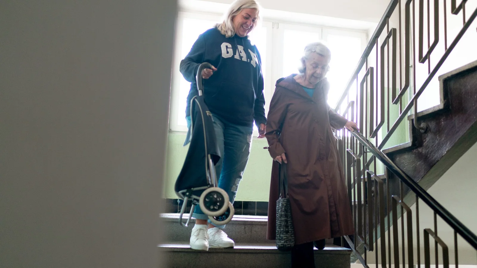 Une accompagnante descend un escalier aux côtés d’une femme âgée équipée d’une aide à la marche